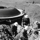 Vintage scene: People in early 20th-century attire view saucer-shaped military aircraft.