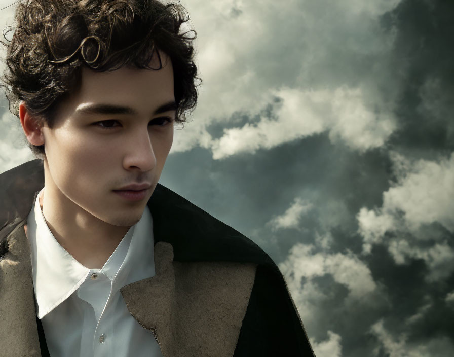 Young man in white shirt and two-toned coat gazes against cloudy sky.