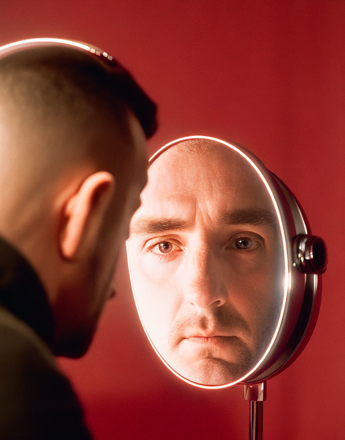 Serious man gazes at reflection in round mirror on red background