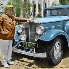 Vintage man in stylish outfit next to classic blue car in elegant garden.