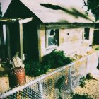 Weathered two-story green house in forest clearing with overgrown foliage and purple flowers