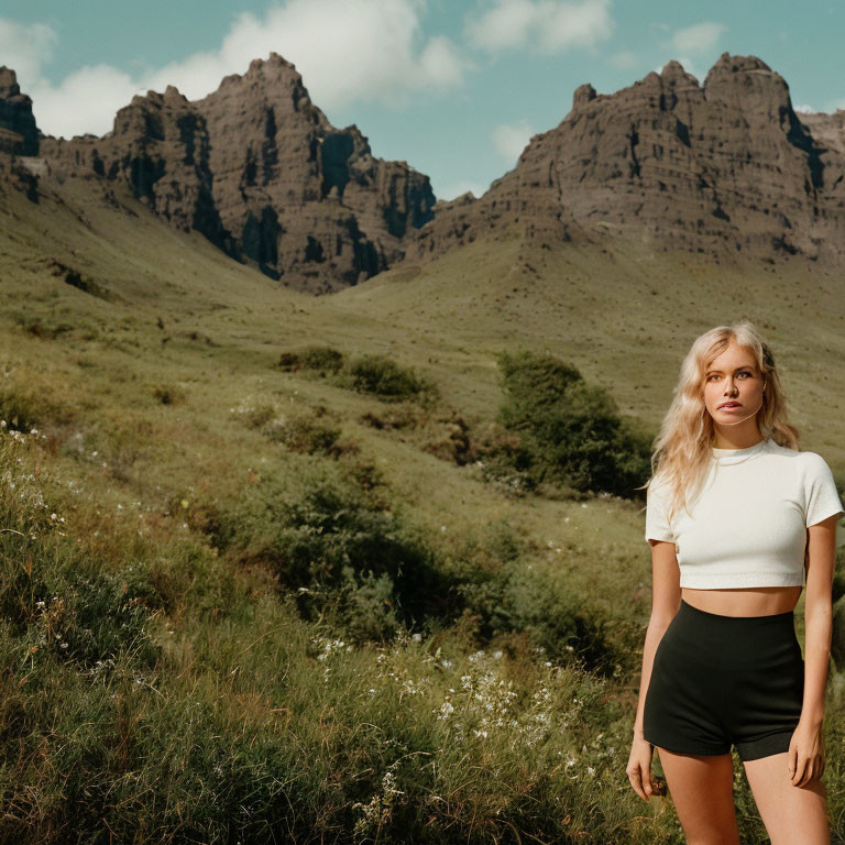 Young woman in white top and black shorts in grassy field with dramatic mountains and cloudy sky.