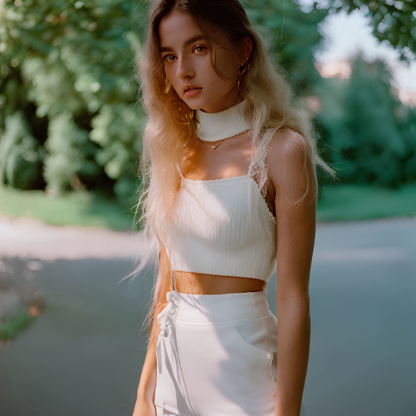 Young Woman in White Crop Top and Skirt Standing on Park Path with Sunlight Filtering Through Trees