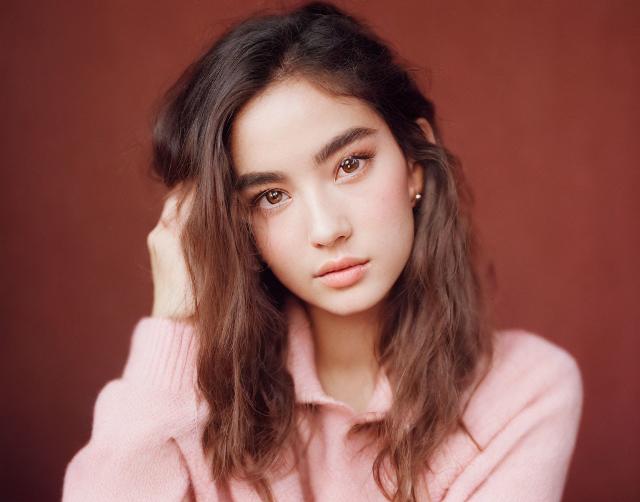 Young woman with wavy brown hair in pink sweater on maroon background