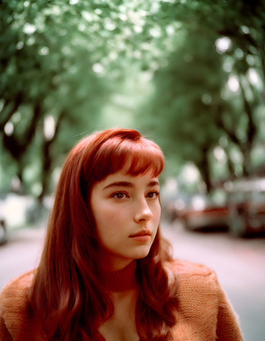 Red-haired woman in peach sweater under green tree canopy on blurred street