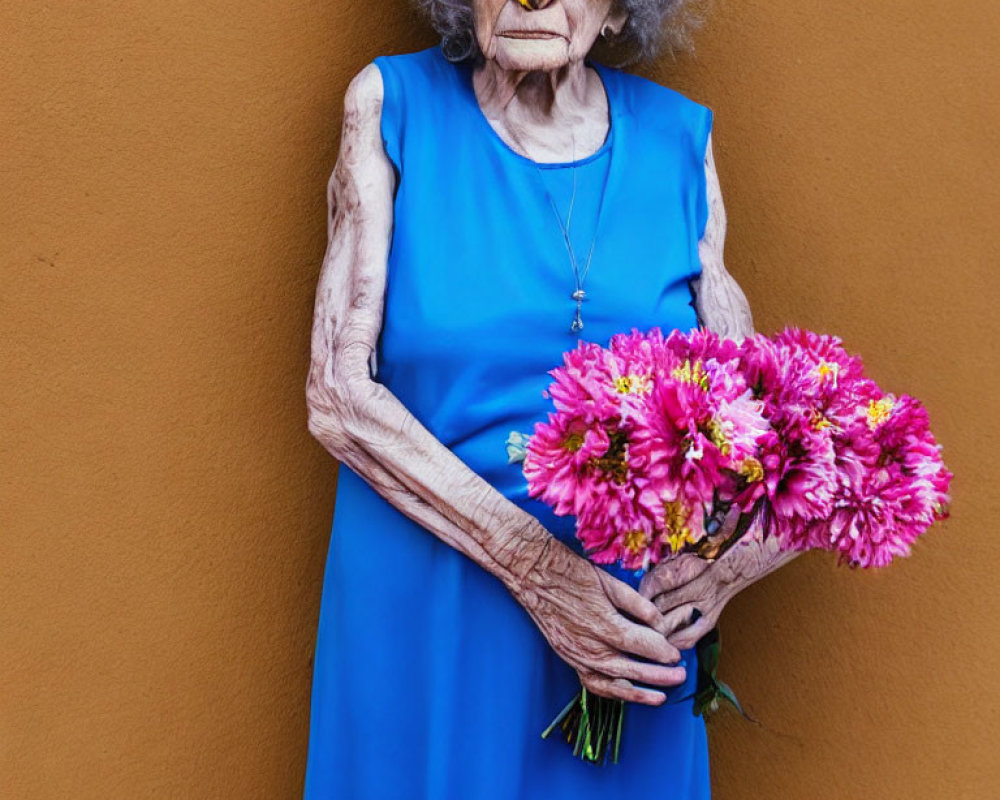 Elderly person in blue dress holding pink flowers against orange wall