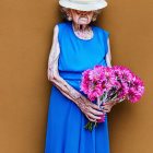 Elderly person in blue dress holding pink flowers against orange wall