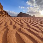 Desert landscape with camels, riders, sand dunes, mountain backdrop, and sunset sky.