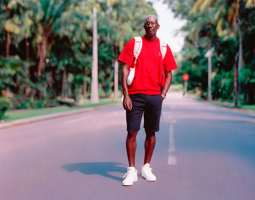 Man in red t-shirt and black shorts on tree-lined road with white satchel and sunglasses