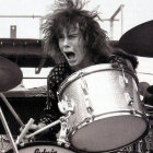 Young drummer with dark hair plays among crowd with eagle-like wings, under bird-filled sky