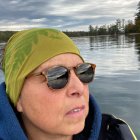 Woman in beanie and sunglasses admires starry night sky over mountains and water.