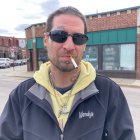 Man with sunglasses and jacket in front of building mural with bleeding wound.