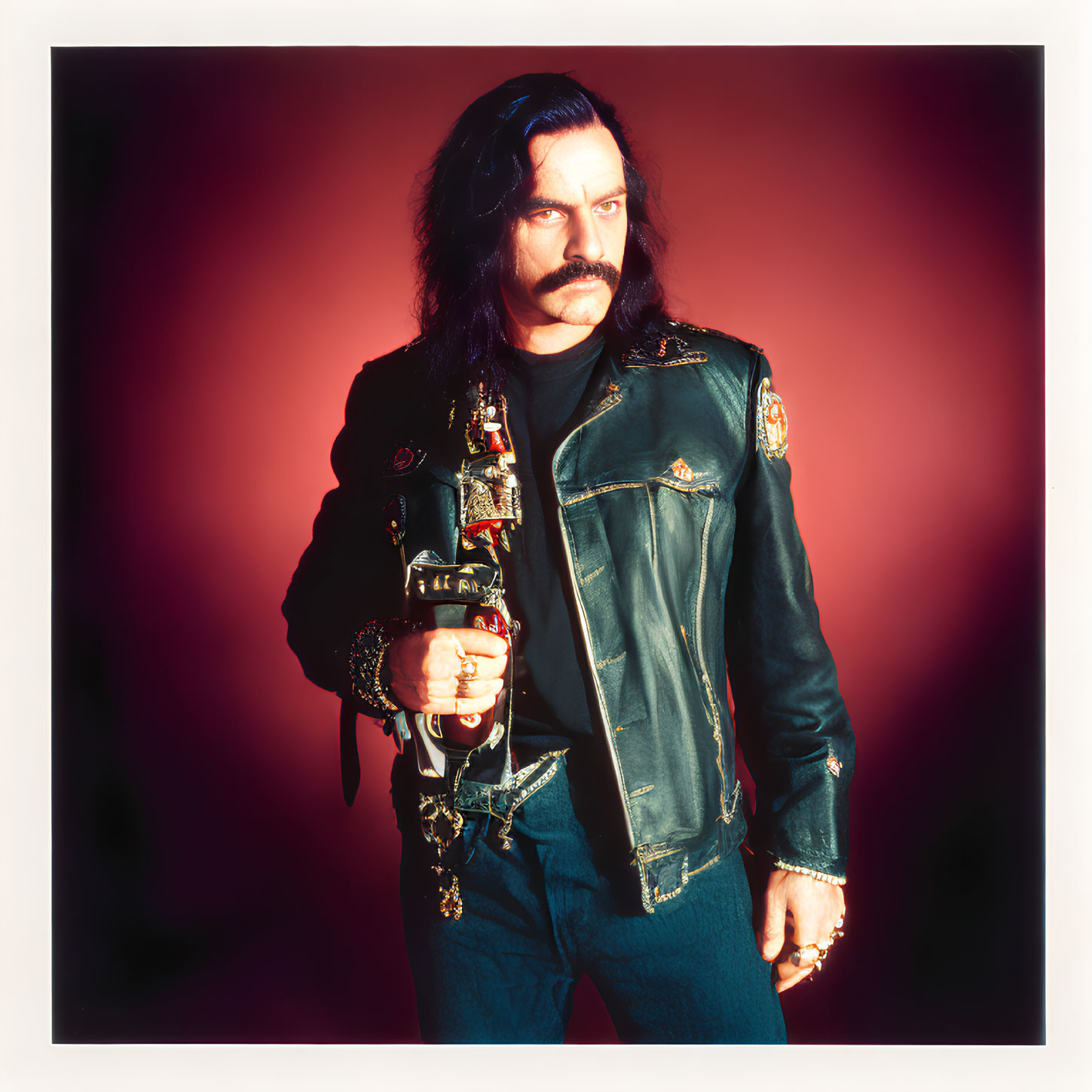 Confident man with dark mustache and long hair holding a bottle in studded leather jacket