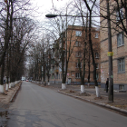 Snowy urban street at dusk with trees, lampposts, cars, and pedestrians