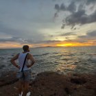 Person admires serene sunset on rocky shore with distant ship and scattered clouds