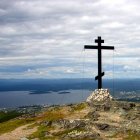 Ornate cross on cliff with wildflowers, calm sea, islands, and cloudy sky