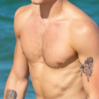 Shirtless male with wavy hair and tattoo on arm, gazing against blue background