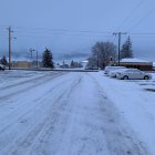 Snow-covered street at dusk with tire tracks, illuminated shops, and mountainous backdrop