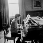 Man in suit plays grand piano in candlelit room with large windows