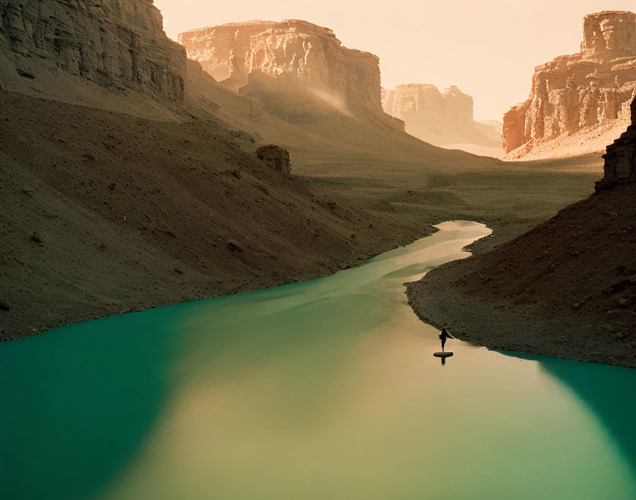 Person standing on narrow rock in turquoise river with brown cliffs and hazy sky