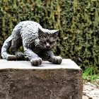 Realistic Grey Striped Cat Sculpture on Wooden Pedestal with Green Hedge Background