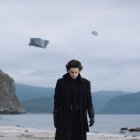 Man in suit and hat gazes at surreal landscape with undulating hills and boat