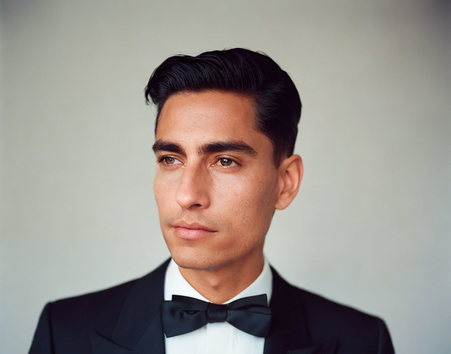 Young man in black tuxedo with slicked-back hair on neutral background