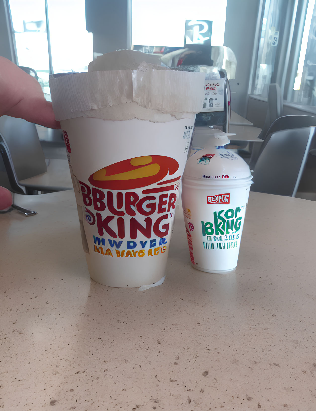 Hand holding Burger King cup with exploded top next to smaller cup on table in bright space