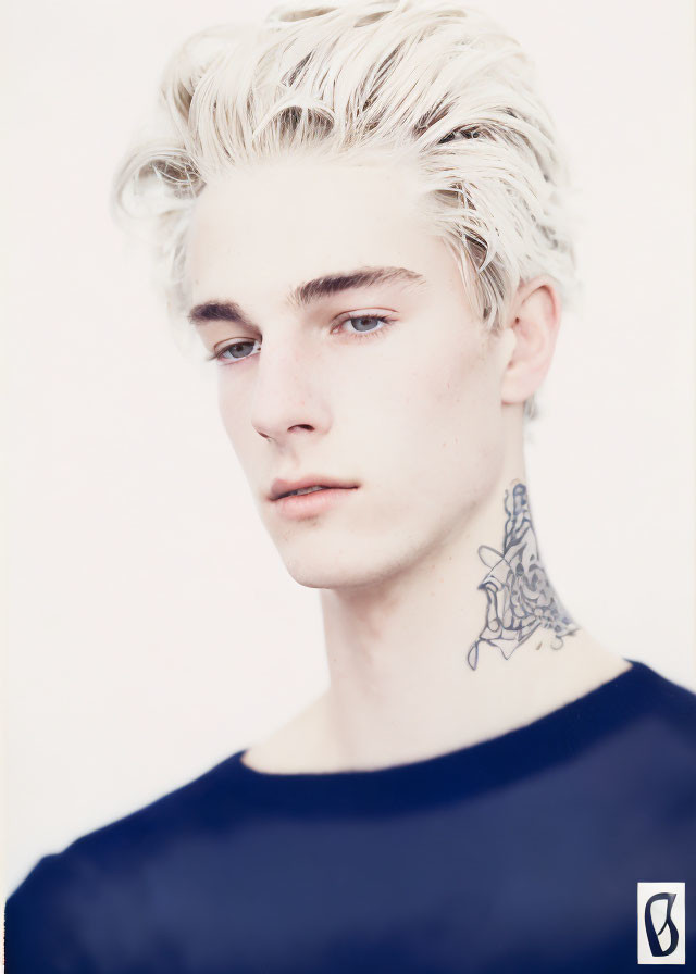 Portrait of young person with platinum blonde hair and floral neck tattoo in dark blue top on pale background.