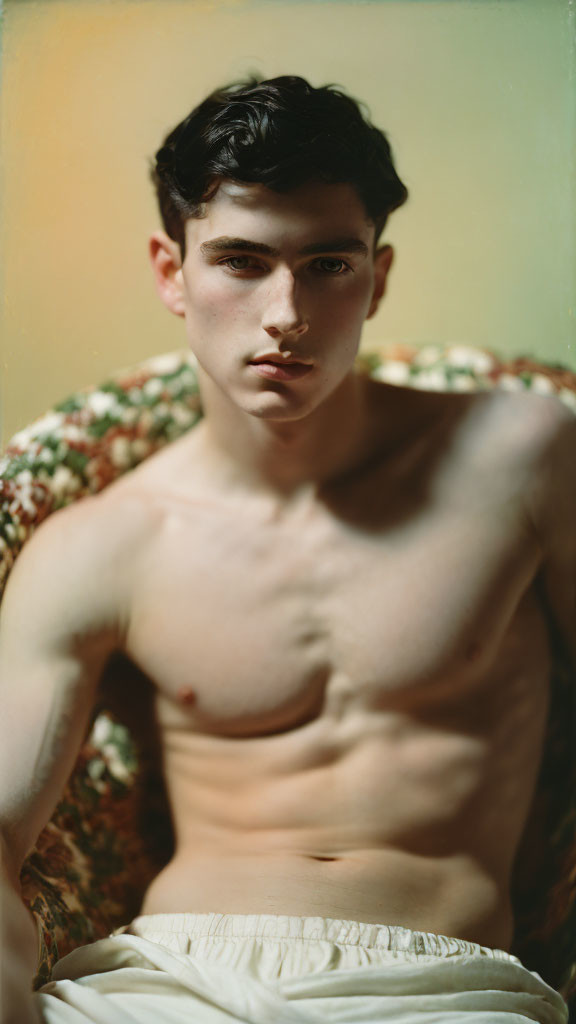 Dark-haired shirtless young man with intense gaze sitting in vintage chair under warm lighting