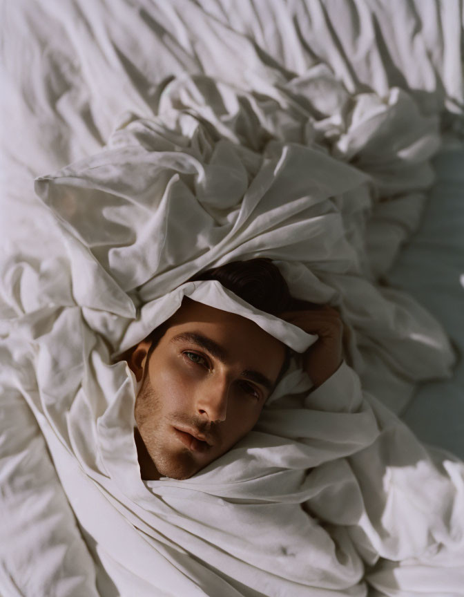 Man in white fabric with hand on forehead in contemplative pose under soft lighting