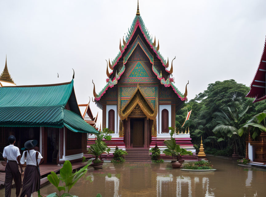 Traditional Thai temple with steep roof and vibrant colors, surrounded by vegetation and two observers