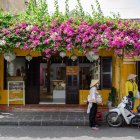 Vintage-style café with pink and purple flowers, warm sunlight, people, and a bicycle.