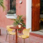 Porcelain cup with lid and spoon on outdoor bistro table near café with green chairs and pink walls