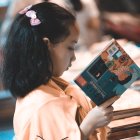 Young girl reading book with pink bow in hair, surrounded by blurred figures