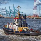 Tugboat sailing in harbor with cranes and industrial structures under partly cloudy sky