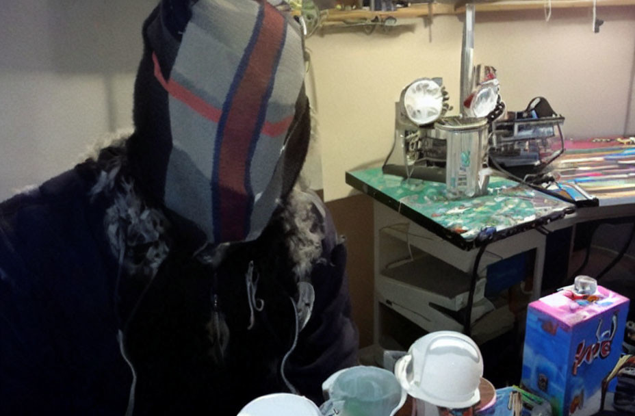 Hooded person at cluttered desk with electronics and cereal box