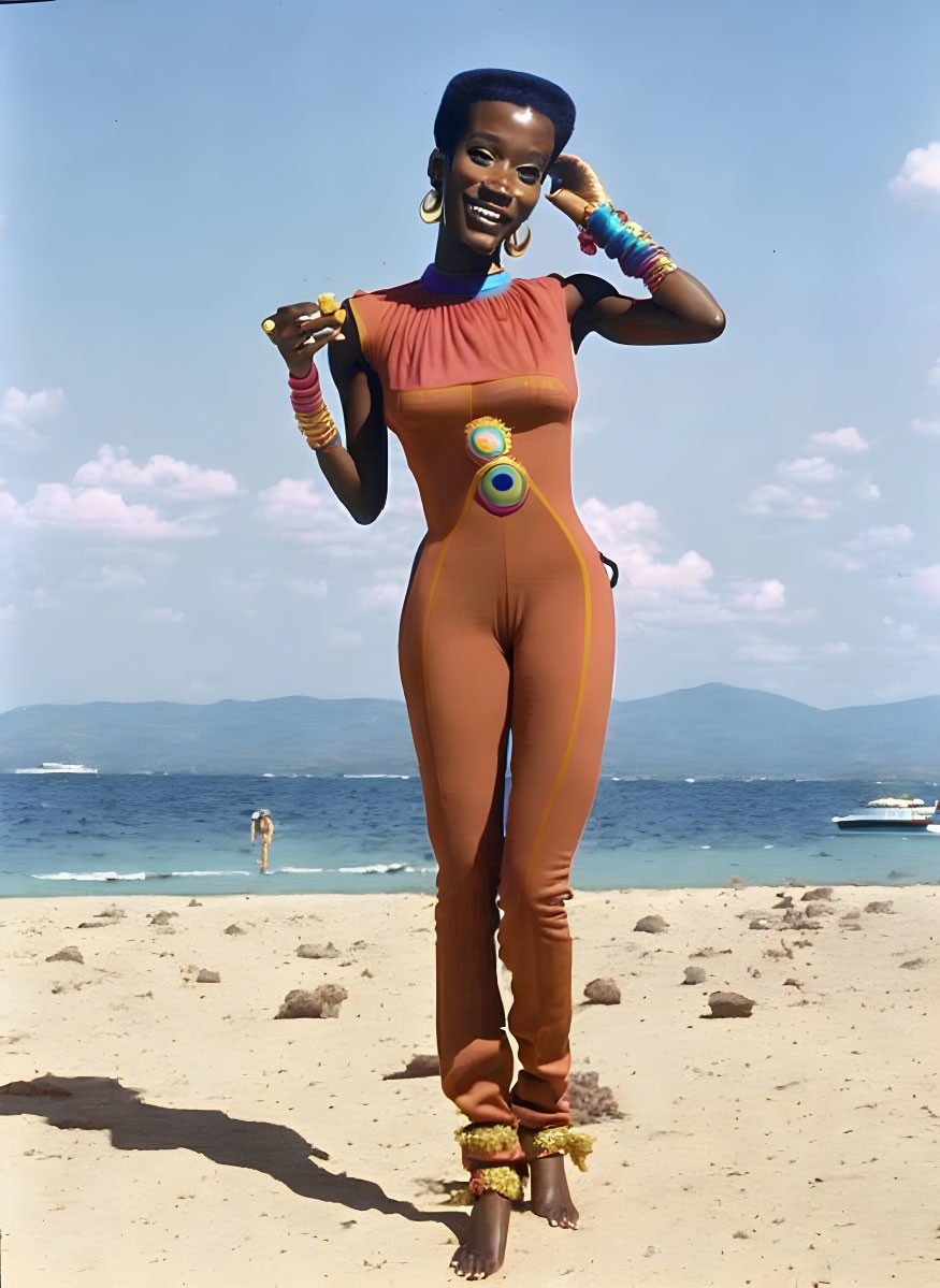 Woman in terracotta jumpsuit on sandy beach with blue water and mountains.