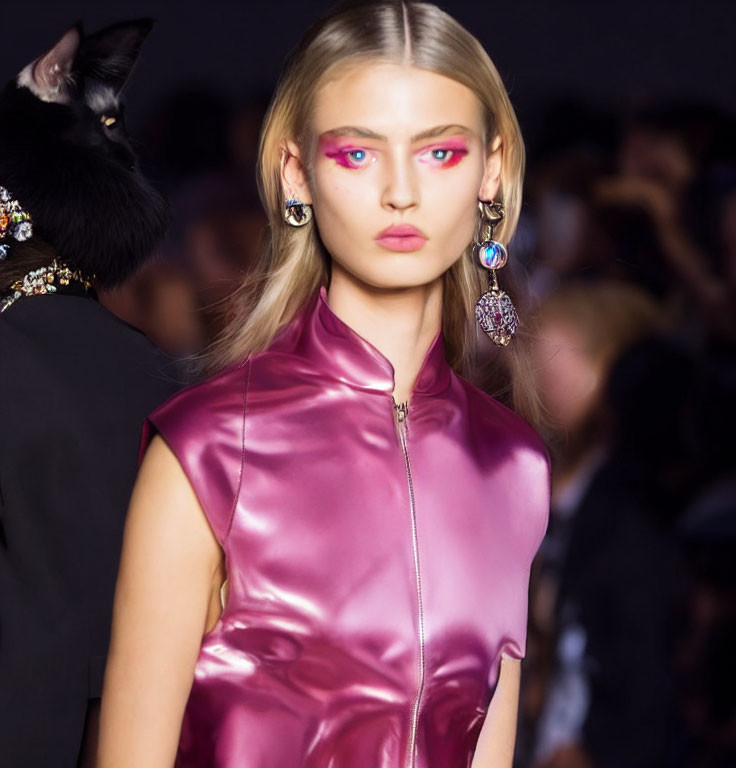 Model with bold pink eye makeup, large earrings, shiny purple top, and black cat.