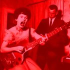 Curly-haired woman plays red bass guitar in red-lit room with man in suit.