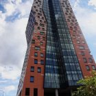 Modern building with orange facade and round windows, green accents, and cylindrical structures against blue sky.