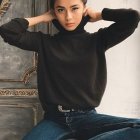 Young woman with curly hair in black top and blue pants, sitting pensively