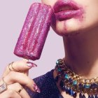 Close-up of woman with purple lipstick biting purple popsicle against pink backdrop