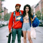 Fashionable couple in urban street style; man with skateboard, red hoodie, and colorful pants, woman