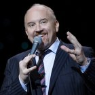 Smiling man with mustache holding microphone in suit against dark background