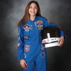 Smiling woman in blue astronaut suit holding helmet, grey backdrop