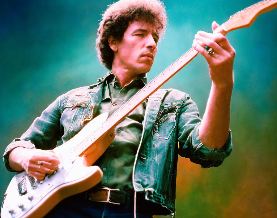 Curly-haired man in denim jacket plays electric guitar under stage lights