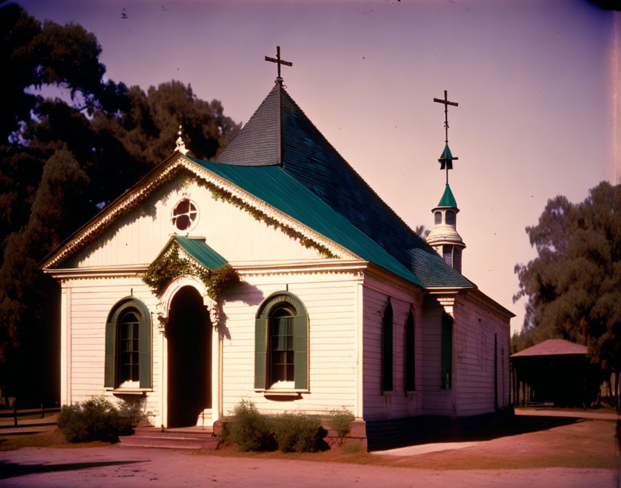 White church with green trim under pink sky, arched windows, cross, lush trees.