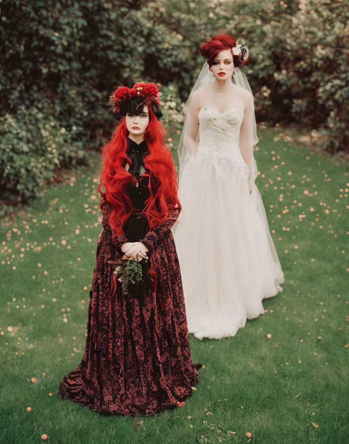 Women in gothic and bridal attire on grass with fallen petals; one in red floral headpiece,