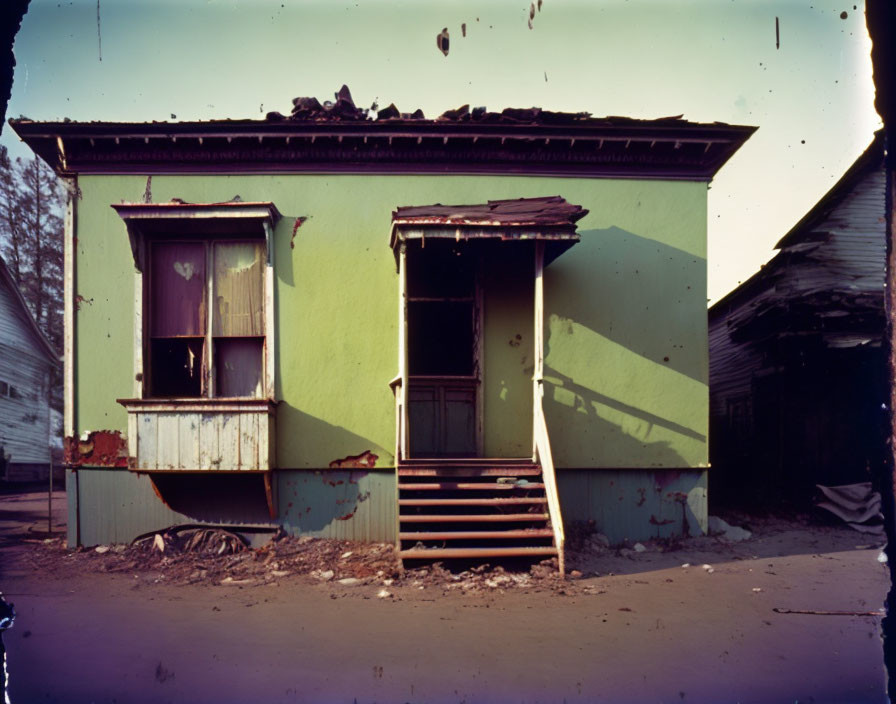 Abandoned green house with damaged roof and stairs, boarded-up window, surrounded by debris under blue sky